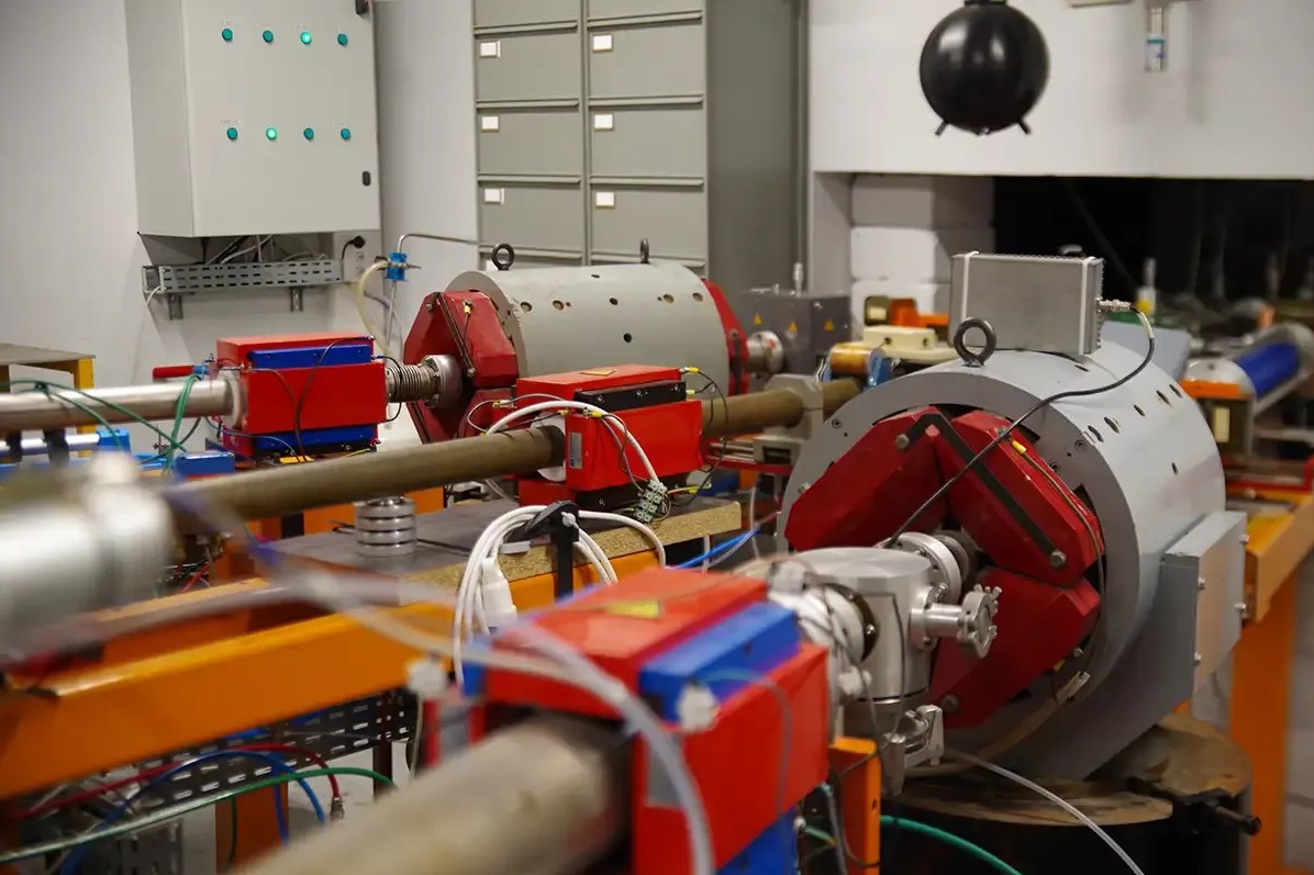 Ligne de faisceau : tube sous vide, quadripôles et dipôles de guidage à bobines rouges, dans un hall d’accélérateur.