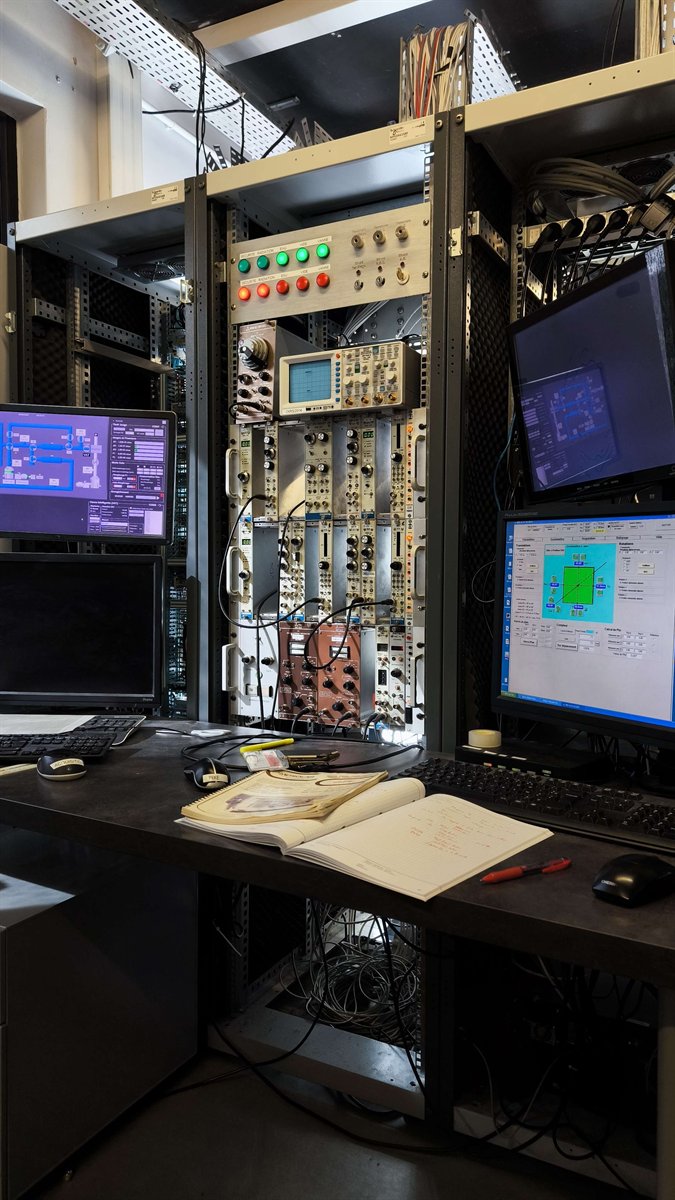 Poste de contrôle : panneau de sécurité à voyants, oscilloscope, châssis de modules d’électronique nucléaire et deux écrans affichant les interfaces historiques du banc.