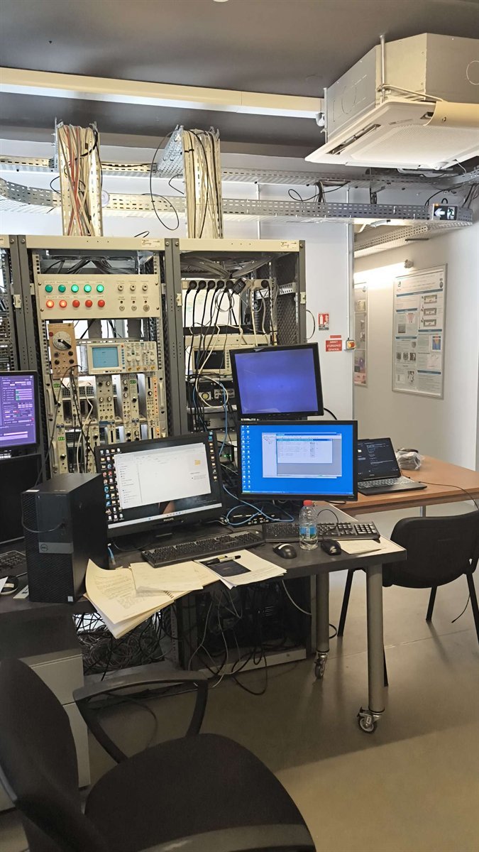 Vue d’ensemble du poste de contrôle : baies d’électronique, écrans, chemins de câbles au plafond.