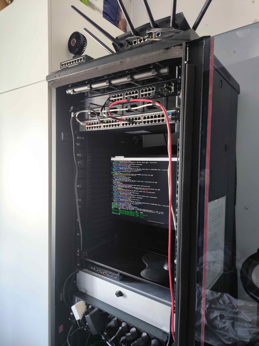 Baie informatique ouverte : commutateurs rackés, routeur Wi-Fi à antennes multiples posé dessus, écran en terminal affichant une configuration réseau, onduleur et multiprise en bas.