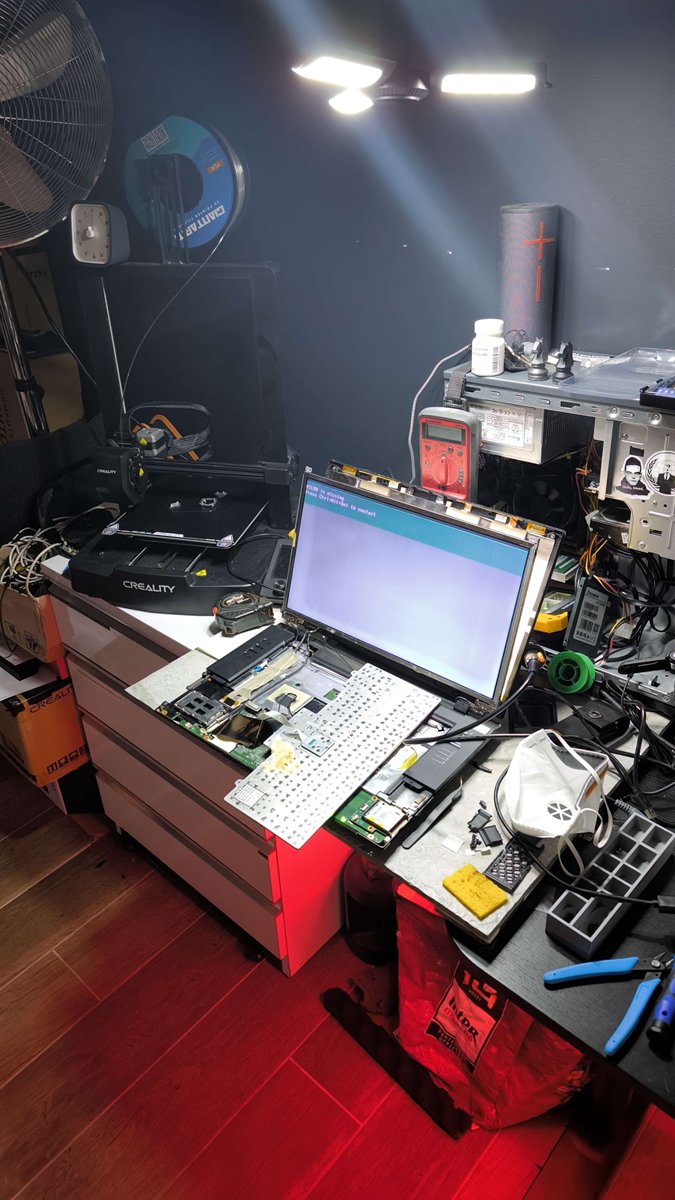 Établi éclairé : ThinkPad ouvert sans coque, écran allumé sur un message de démarrage, outils et imprimante 3D autour.